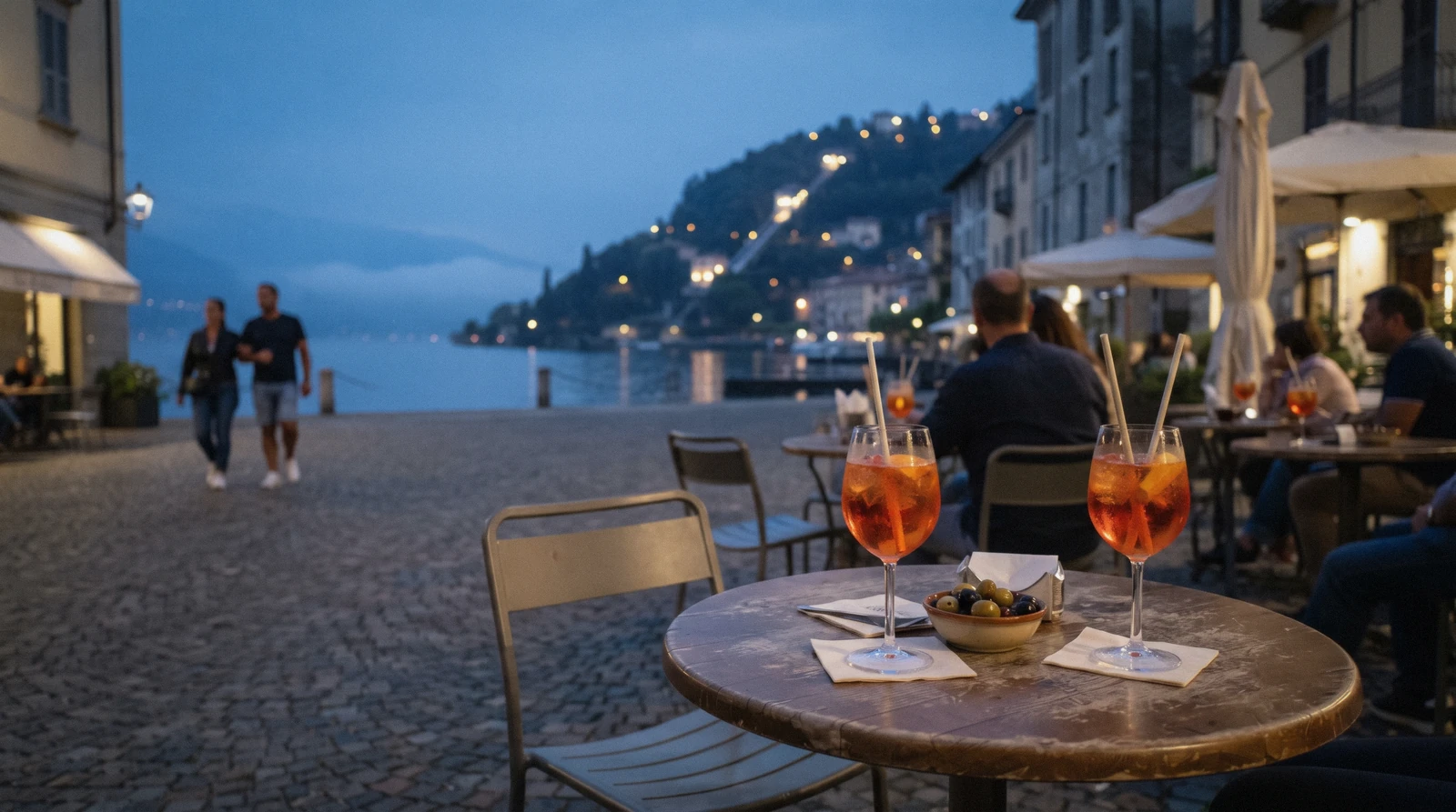 Two spritz glasses on an outdoor table on Piazza Cavour at blue hour with the lit Brunate hillside across the lake, the aperitivo scene of bars in Como